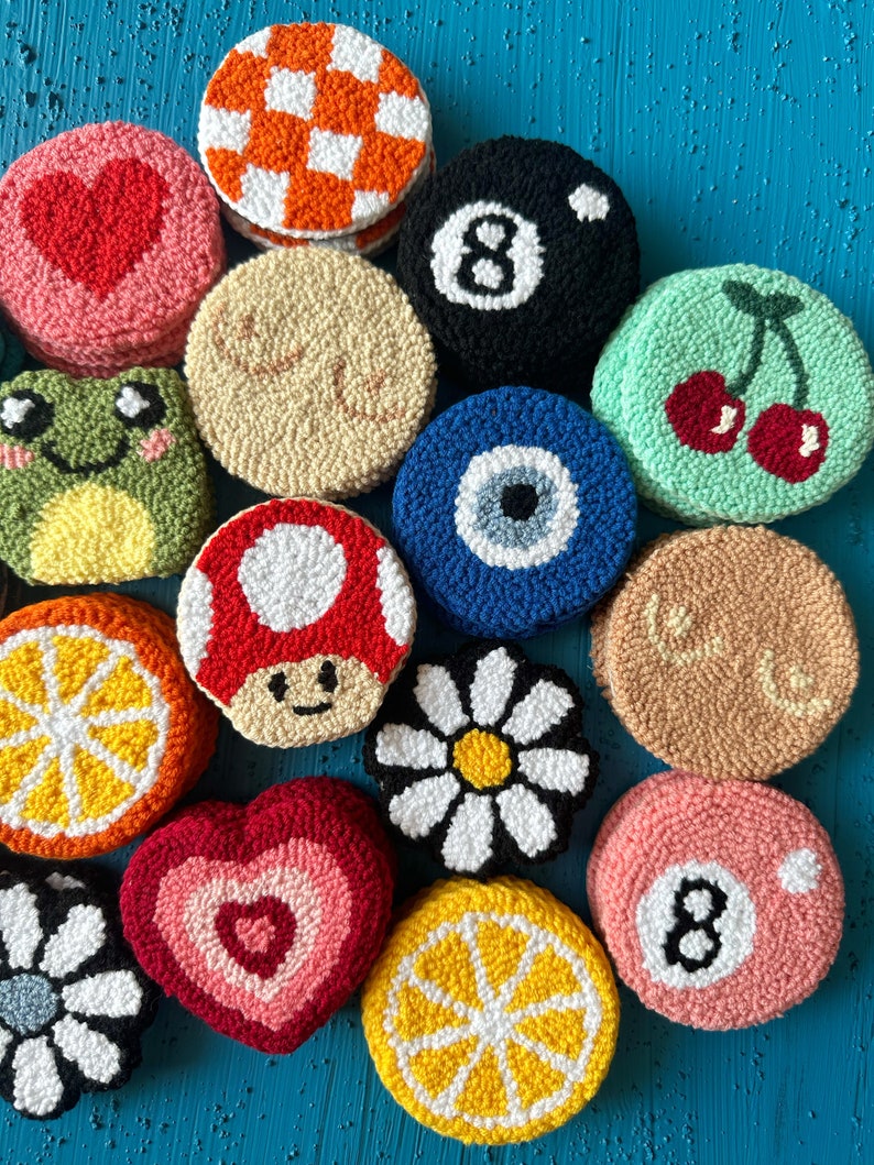 May include: A collection of 12 colorful, round, crocheted coasters with various designs, including a heart, a mushroom, a daisy, a lemon, a cherry, an eye, a frog, a checkered pattern, and the number 8.