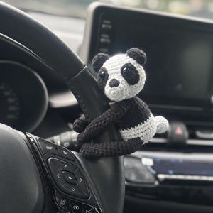以下が含まれることがあります： 車のステアリングホイールにぶら下がっている、かぎ針編みで作られたパンダのぬいぐるみ。パンダは白黒で、顔は白く、目は黒いです。ステアリングホイールには、車の機能を制御するためのボタンがあります。