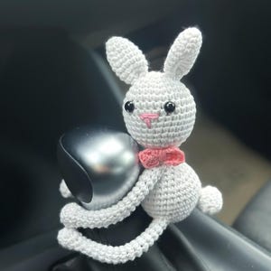 Könnte beinhalten: Ein weißer Häkelhase mit rosa Ohren und einer rosa Schleife. Der Hase sitzt auf dem Schalthebel eines Autos.