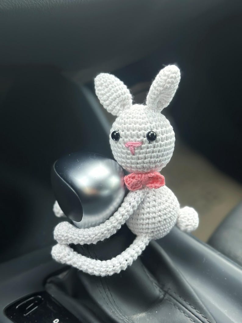 K&ouml;nnte beinhalten: Ein wei&szlig;er H&auml;kelhase mit rosa Ohren und einer rosa Schleife. Der Hase sitzt auf dem Schalthebel eines Autos.