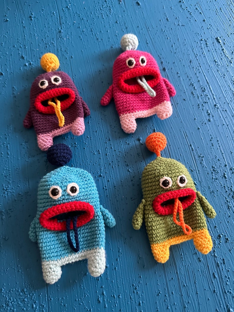 Puede incluir: Cuatro juguetes de peluche de crochet con forma de monstruos con bocas grandes y ojos saltones. Los monstruos son de diferentes colores: morado, rosa, azul y verde.