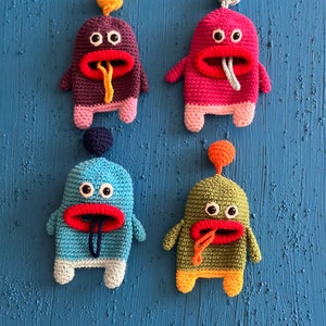 Puede incluir: Cuatro juguetes de peluche de crochet con forma de monstruos con cuerpos y caras de diferentes colores. Los monstruos tienen ojos grandes, bocas rojas y lenguas largas que sobresalen. Los juguetes est&aacute;n dispuestos en fila sobre un fondo azul.