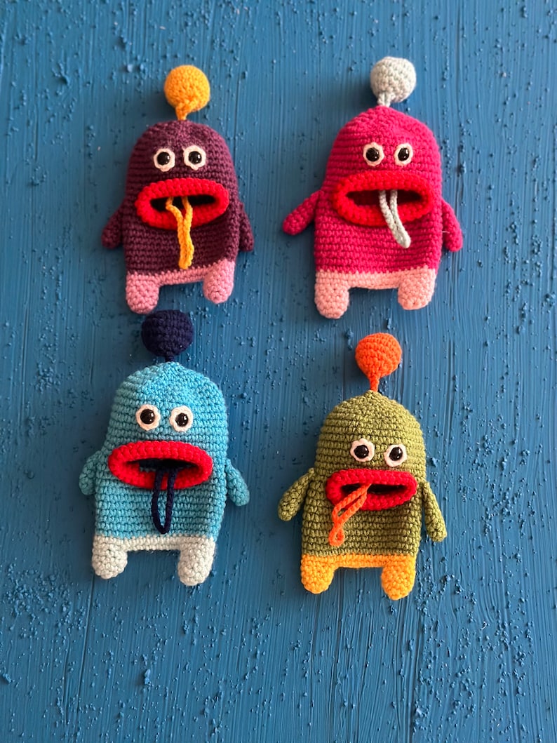 Puede incluir: Cuatro juguetes de peluche de crochet con forma de monstruos con cuerpos y caras de diferentes colores. Los monstruos tienen ojos grandes, bocas rojas y lenguas largas que sobresalen. Los juguetes est&aacute;n dispuestos en fila sobre un fondo azul.