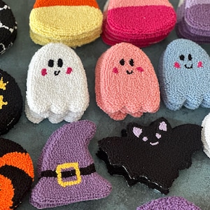 May include: A collection of colorful, textured Halloween-themed decorations. The assortment includes candy corn, ghosts, a witch's hat, and bats. The decorations are made of a soft, looped material and feature embroidered details.