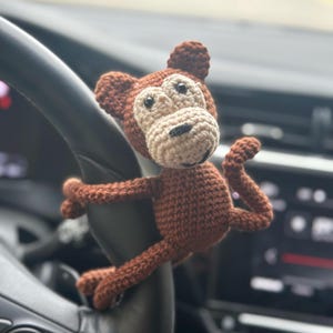 Könnte beinhalten: Ein braunes Häkel-Affenspielzeug mit beigefarbenem Gesicht und schwarzen Augen hängt am Lenkrad eines Autos.