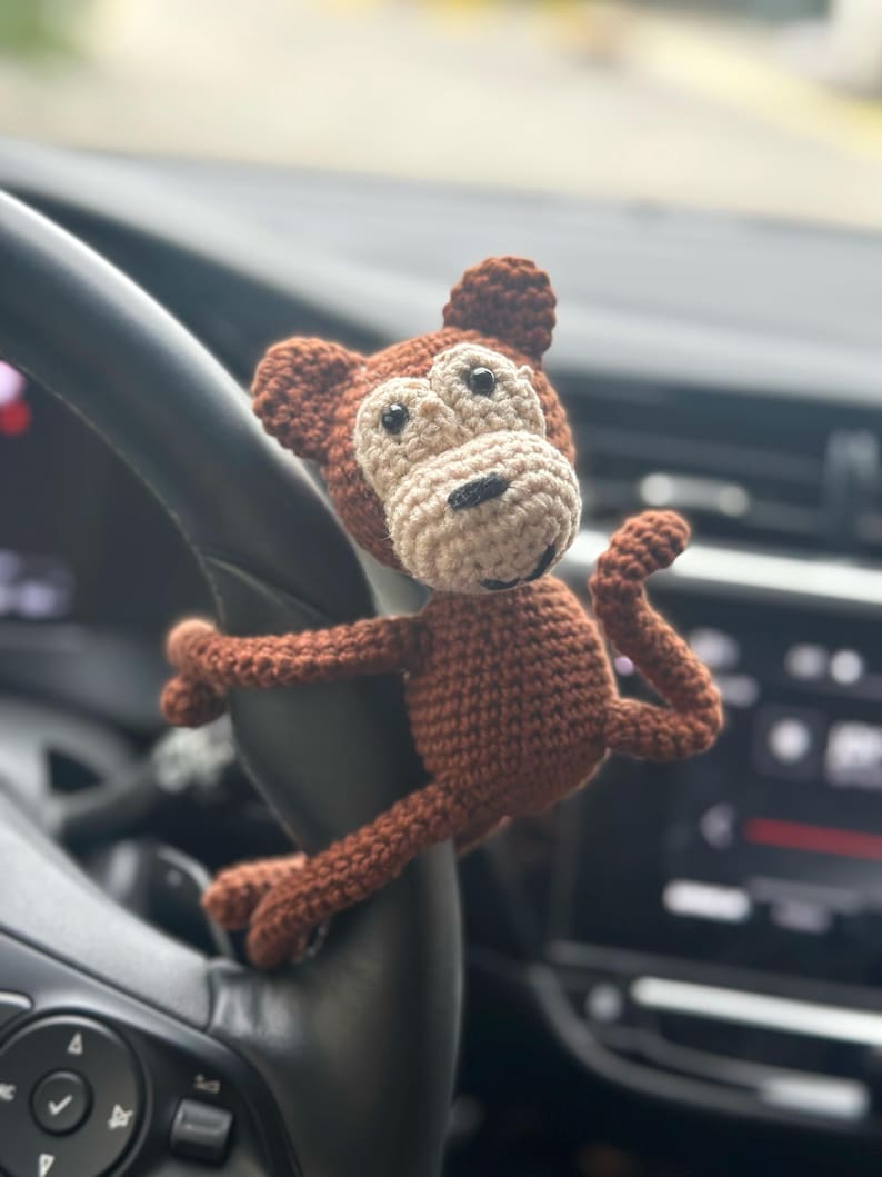 K&ouml;nnte beinhalten: Ein braunes H&auml;kel-Affenspielzeug mit beigefarbenem Gesicht und schwarzen Augen h&auml;ngt am Lenkrad eines Autos.