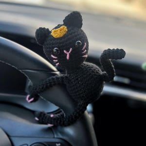 May include: A black crochet cat with pink whiskers and accents, and a yellow crown. The handmade cat is perched on a black steering wheel. The cat's eyes are large and round, giving it a cute appearance.