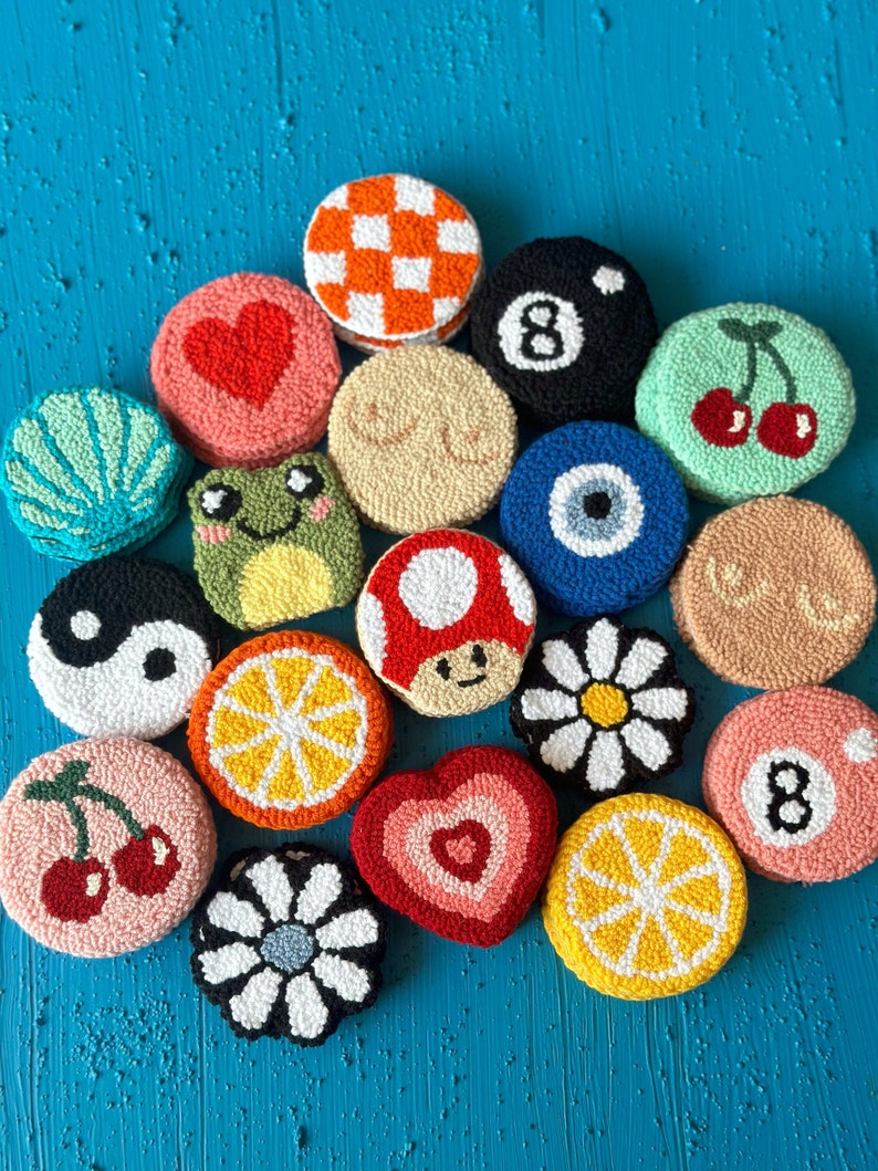 May include: A collection of 16 colorful, round, hand-stitched felt ornaments. The ornaments feature various designs, including a mushroom, a cherry, a daisy, a lemon, a heart, a frog, a yin-yang symbol, an evil eye, a seashell, a checkered pattern, a pool ball, and a number 8.