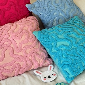 May include: Four decorative throw pillows with a unique, textured, wavy design. The pillows are in shades of pink, blue, and turquoise. There are two small, embroidered decorations on the white fabric: a white bunny and a green seashell.