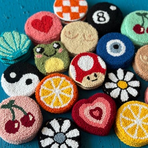 May include: A collection of 15 colorful, round, hand-stitched felt ornaments with various designs, including a heart, a mushroom, a daisy, a cherry, an orange slice, a frog, a yin-yang symbol, an evil eye, a checkered pattern, a pool ball, and a breast shape.