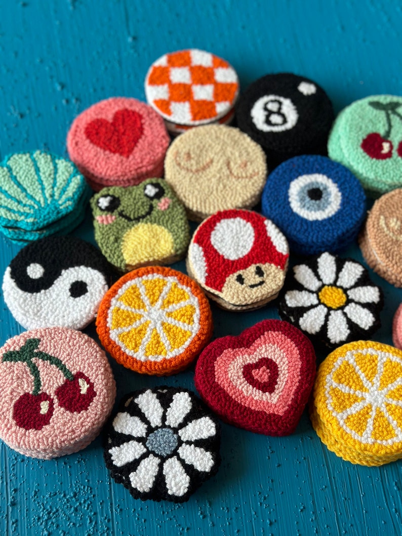 May include: A collection of 15 colorful, round, hand-stitched felt ornaments with various designs, including a heart, a mushroom, a daisy, a cherry, an orange slice, a frog, a yin-yang symbol, an evil eye, a checkered pattern, a pool ball, and a breast shape.
