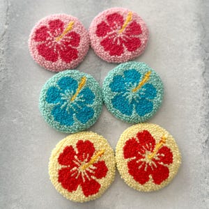 May include: Six round, colorful, felt flower coasters with a hibiscus design. The coasters are in pink, blue, red, and yellow.