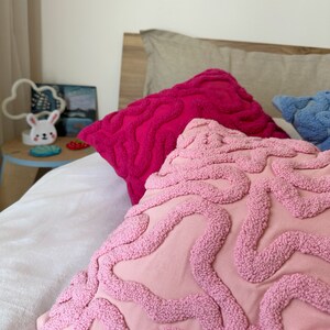 May include: Pink and fuchsia throw pillows with a textured, wavy design. The pillows are on a white bed with a beige pillow in the background.