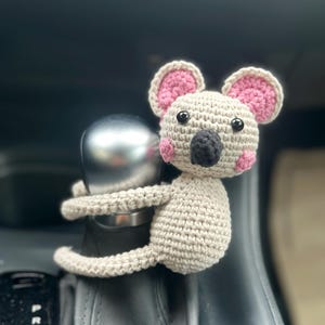May include: A crocheted koala toy with pink ears and a black nose, sitting on a car's gear stick.