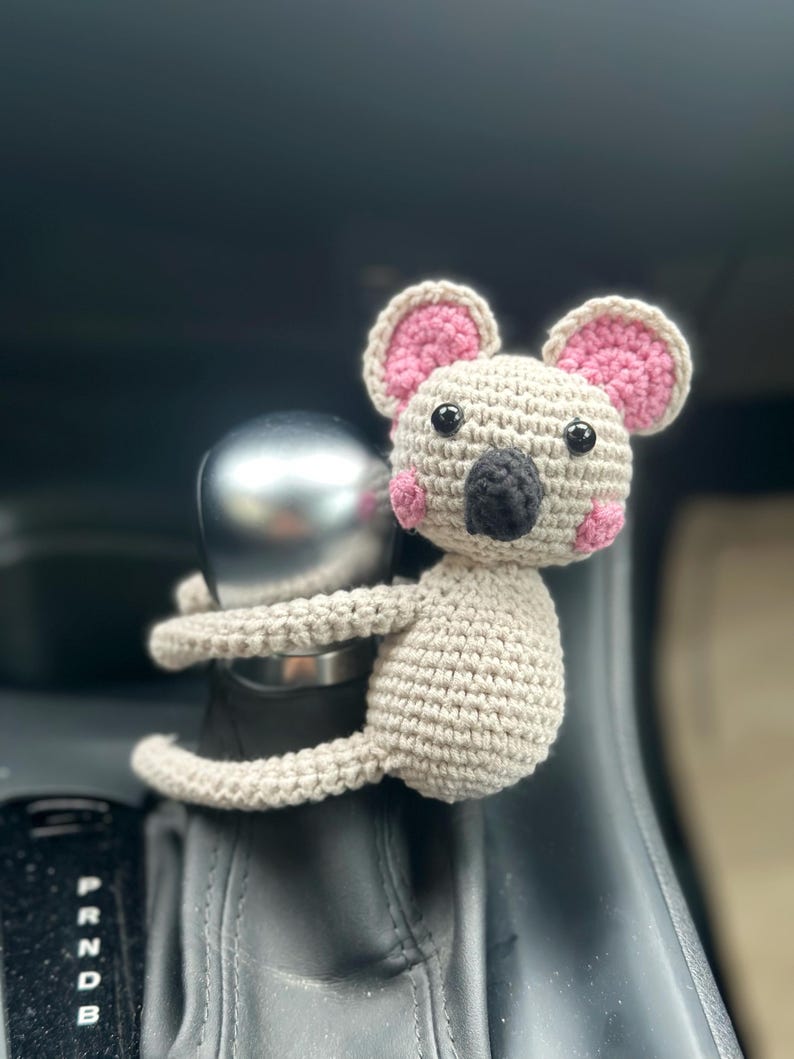 K&ouml;nnte beinhalten: Ein geh&auml;keltes Koala-Spielzeug mit rosa Ohren und einer schwarzen Nase, das auf dem Schalthebel eines Autos sitzt.