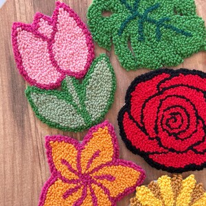 May include: A collection of colorful, handmade floral coasters. The set includes a pink tulip, a green leaf, a red rose, an orange and pink flower, and a yellow sunflower. Each coaster is made with a textured, looped yarn design.