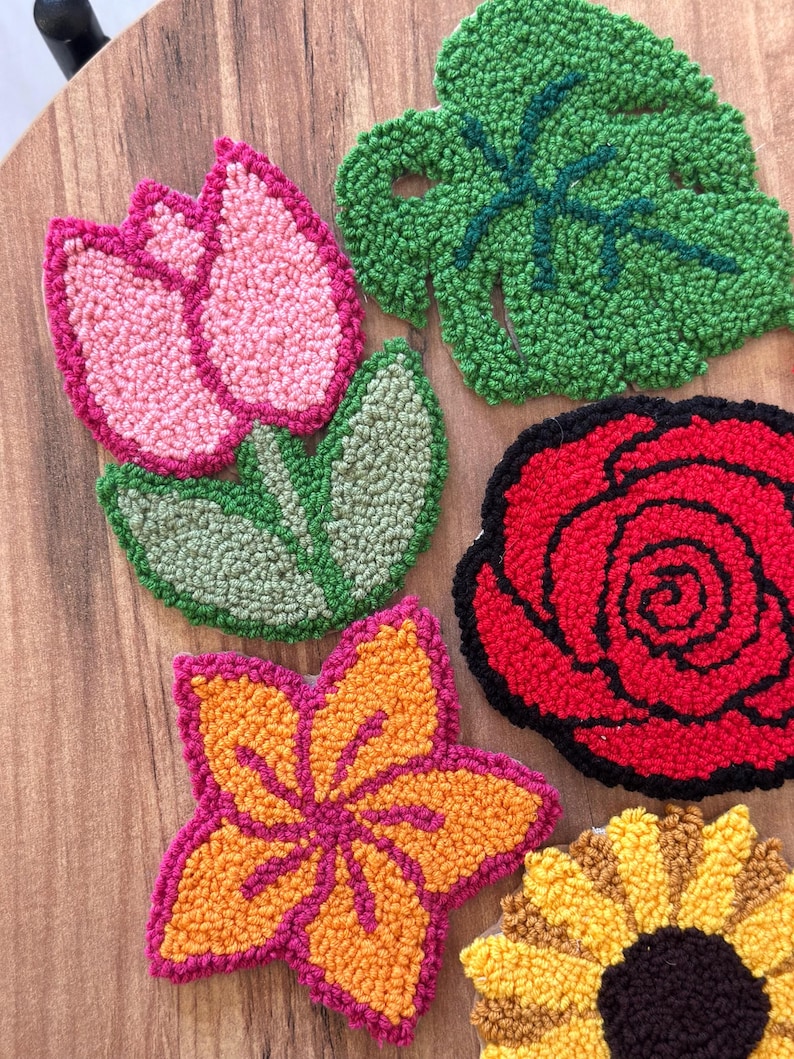 May include: A collection of colorful, handmade floral coasters. The set includes a pink tulip, a green leaf, a red rose, an orange and pink flower, and a yellow sunflower. Each coaster is made with a textured, looped yarn design.