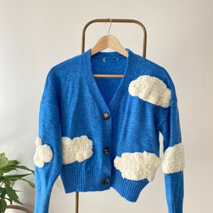 May include: A blue cardigan with white fluffy cloud appliques. The cardigan has a v-neck and button closure.
