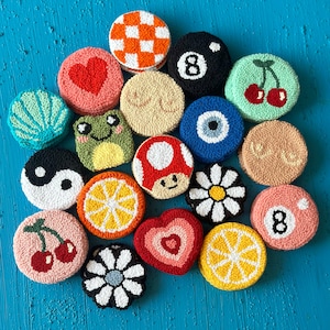 May include: A collection of 17 colorful, round, embroidered felt ornaments with various designs, including cherries, a mushroom, a heart, a lemon, a daisy, a frog, an evil eye, a yin yang symbol, a checkered pattern, a pool ball, and the number 8.