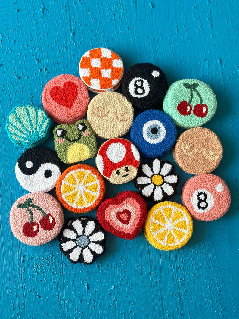 May include: A collection of 17 colorful, round, embroidered felt ornaments with various designs, including cherries, a mushroom, a heart, a lemon, a daisy, a frog, an evil eye, a yin yang symbol, a checkered pattern, a pool ball, and the number 8.