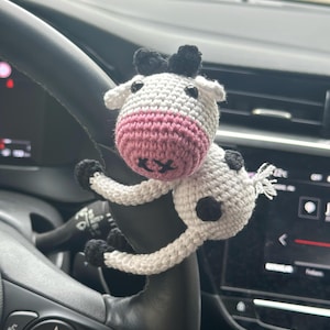 K&ouml;nnte beinhalten: Eine geh&auml;kelte wei&szlig;e Kuh mit schwarzen Flecken und rosa Ohren, die an einem Autolenkrad h&auml;ngt.