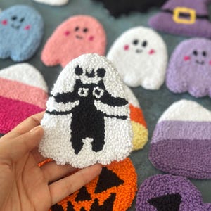 May include: A collection of handmade Halloween-themed plush decorations. The assortment includes ghosts, a skull, a bat, a witch's hat, a jack-o'-lantern, and candy corn. The plushies are made with various colors, including white, orange, purple, pink, and blue.