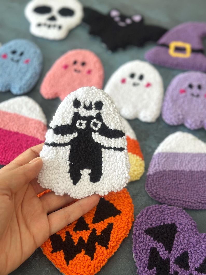 May include: A collection of handmade Halloween-themed plush decorations. The assortment includes ghosts, a skull, a bat, a witch's hat, a jack-o'-lantern, and candy corn. The plushies are made with various colors, including white, orange, purple, pink, and blue.