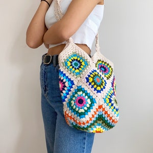 May include: A colorful crocheted tote bag with a white background and a variety of bright colors, including blue, green, yellow, orange, pink, and red. The bag has a long strap and is worn over the shoulder.