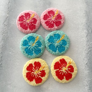 May include: Six round, colorful, felt-covered coasters with a hibiscus flower design. The coasters are in pink, blue, yellow, and red.
