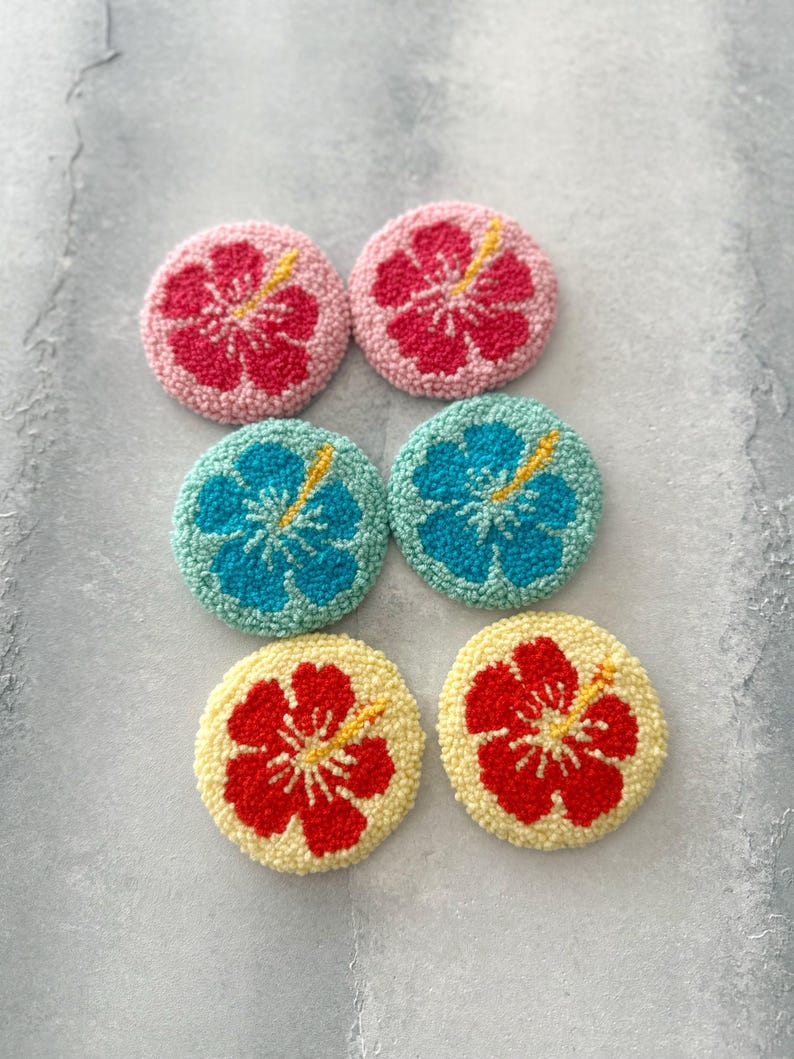 May include: Six round, colorful, felt-covered coasters with a hibiscus flower design. The coasters are in pink, blue, yellow, and red.