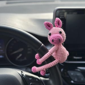 May include: A pink crocheted pig, designed as a car accessory. The pig has a light pink body, darker pink snout and ears, and black eyes. It is attached to a black steering wheel. A charming, handmade decorative item.