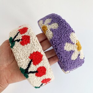 Pu&ograve; includere: Due fasce per capelli fatte a mano, una con sfondo bianco e ciliegie rosse, l'altra con sfondo viola e margherite bianche.