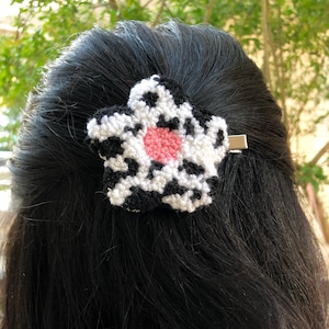 Może przedstawiać: Spinka do włosów w kształcie kwiatu z czarno-białym wzorem i różowym środkiem. Spinka jest przymocowana do srebrnej metalowej spinki. Akcesorium do włosów jest umieszczone na tle ciemnych włosów.