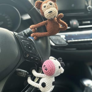 K&ouml;nnte beinhalten: Ein brauner geh&auml;kelter Affe und eine wei&szlig;e geh&auml;kelte Kuh mit schwarzen Flecken und rosa Ohren, die an einem Autolenkrad h&auml;ngen.