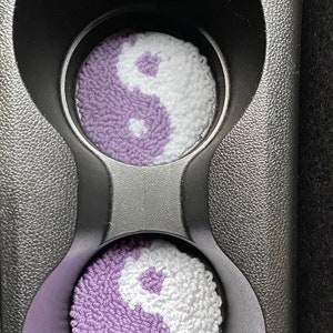 May include: Two purple and white yin yang designs are in the cup holders of a car. The designs are made of yarn and are circular.