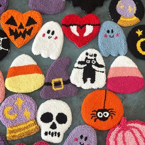 May include: A collection of colorful, handmade Halloween-themed coasters. Designs include a jack-o'-lantern, ghost, bat, spider, skull, witch hat, and candy corn. The coasters are made with a textured, looped yarn technique.