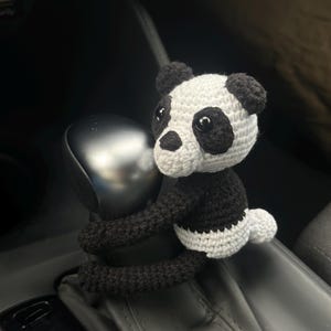 以下が含まれることがあります： 黒と白の毛糸で編まれたパンダのぬいぐるみが、車のシフトレバーに取り付けられています。パンダは腕でレバーを握っており、かわいい表情をしています。