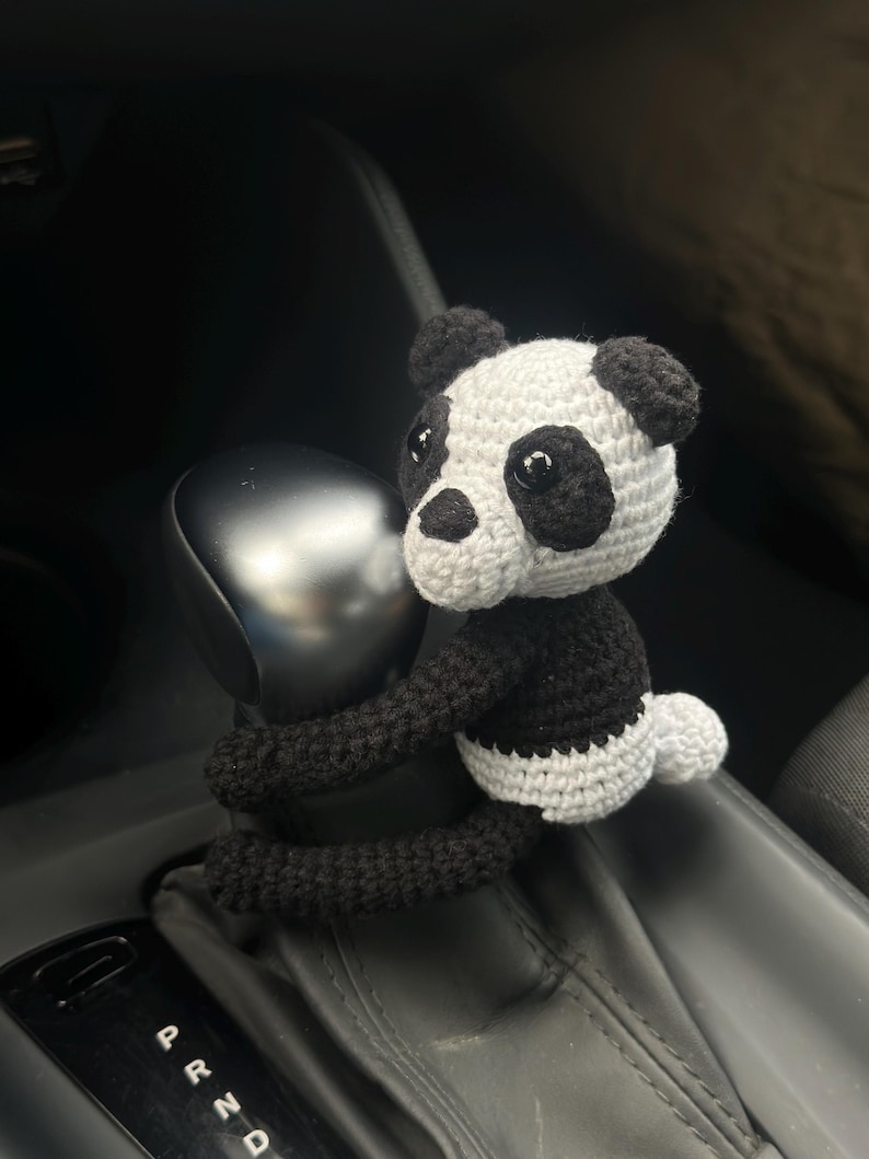 以下が含まれることがあります： 黒と白の毛糸で編まれたパンダのぬいぐるみが、車のシフトレバーに取り付けられています。パンダは腕でレバーを握っており、かわいい表情をしています。