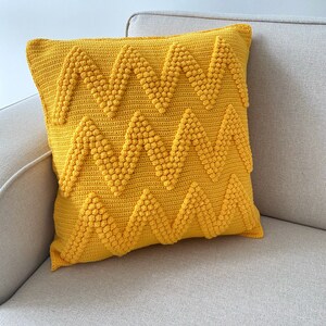May include: A yellow crocheted cushion with a chevron pattern. The cushion is made with a textured yarn that creates a bumpy surface.
