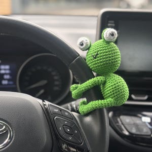 以下が含まれることがあります： 白い目をした緑色の編みぐるみカエルのおもちゃが車のステアリングホイールにぶら下がっています。ステアリングホイールは黒い革製で、「TRIP」と「AUDIO」と書かれたボタンが付いています。