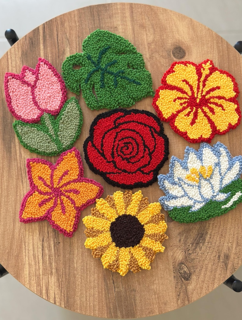 May include: A collection of colorful, handmade floral coasters. The set includes a pink tulip, a green leaf, a red rose, a yellow hibiscus, an orange flower, a yellow sunflower, and a blue and white water lily. The coasters are arranged on a wooden surface.