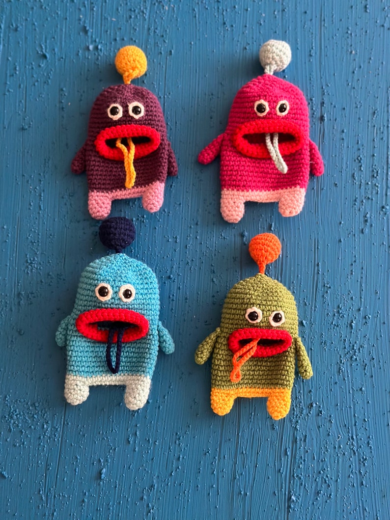Puede incluir: Cuatro juguetes de peluche de crochet con forma de monstruos con cuerpos y caras de diferentes colores. Los monstruos tienen ojos grandes, bocas rojas y lenguas largas que sobresalen. Los juguetes est&aacute;n dispuestos en fila sobre un fondo azul.