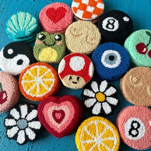 May include: A collection of colorful, round, hand-stitched felt ornaments with various designs, including a heart, a frog, a lemon slice, a daisy, a mushroom, a yin-yang symbol, an evil eye, a cherry, a checkered pattern, a pool ball, and a number eight.