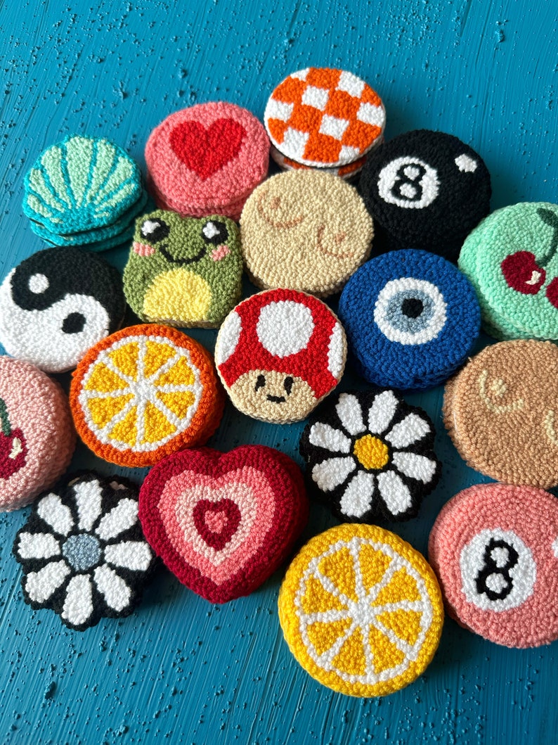 May include: A collection of colorful, round, hand-stitched felt ornaments with various designs, including a heart, a frog, a lemon slice, a daisy, a mushroom, a yin-yang symbol, an evil eye, a cherry, a checkered pattern, a pool ball, and a number eight.