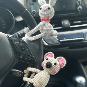 K&ouml;nnte beinhalten: Zwei geh&auml;kelte Stofftiere h&auml;ngen am Lenkrad eines Autos. Der wei&szlig;e Hase hat rosa Ohren und eine rosa Schleife. Der beige Koala hat rosa Ohren und eine schwarze Nase.