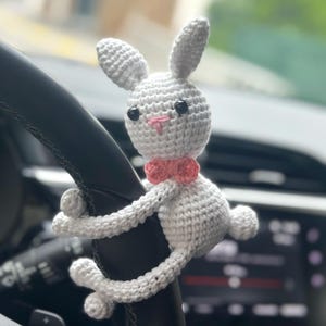 K&ouml;nnte beinhalten: Ein wei&szlig;er H&auml;kelhase mit rosa Akzenten h&auml;ngt am Lenkrad eines Autos. Der Hase hat schwarze Augen und eine rosa Nase.