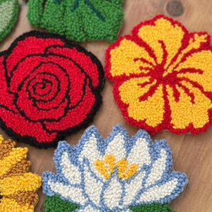 May include: A collection of colorful, textured, floral-shaped coasters. Designs include a red rose, yellow hibiscus, blue and white water lily, and a green leaf. The coasters are made with a looped yarn technique.
