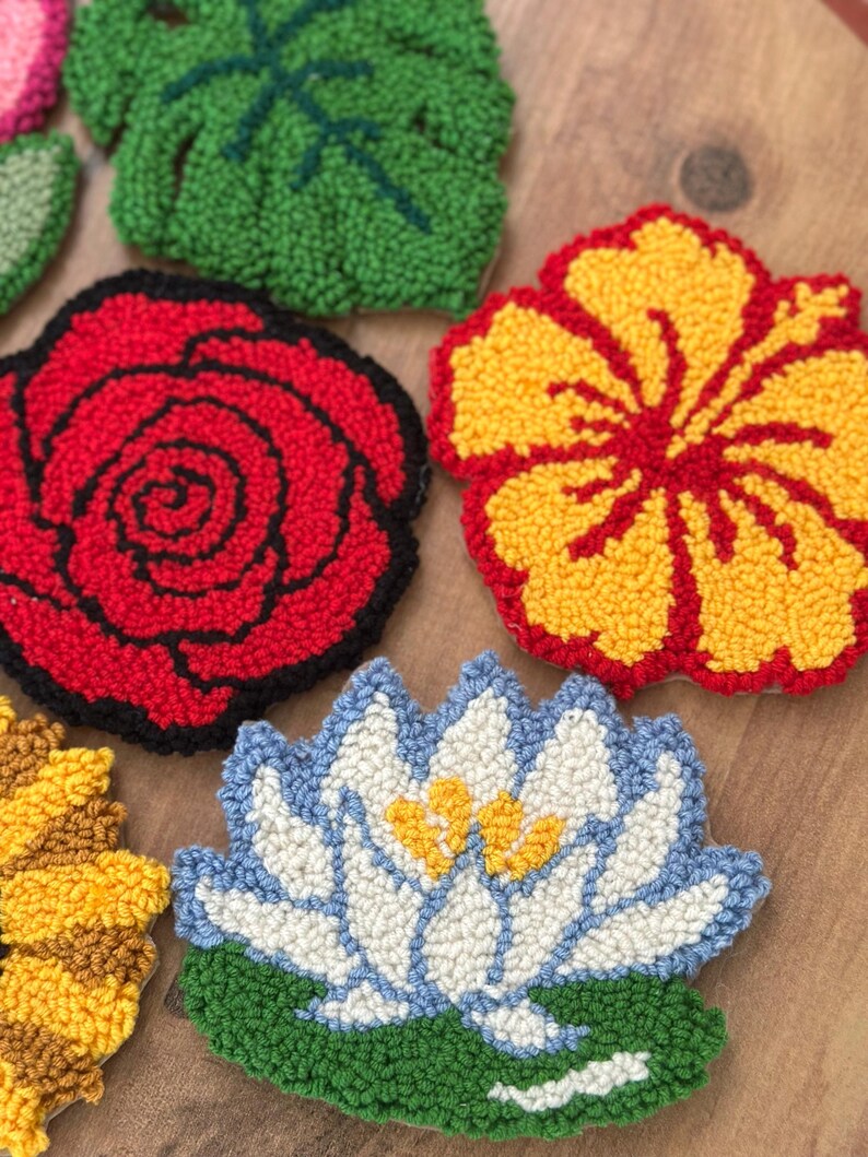 May include: A collection of colorful, textured, floral-shaped coasters. Designs include a red rose, yellow hibiscus, blue and white water lily, and a green leaf. The coasters are made with a looped yarn technique.