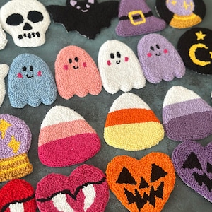 May include: A collection of handmade Halloween-themed coasters. Designs include a white skull, a black bat, a purple witch hat, ghosts, candy corn, and jack-o'-lanterns. The coasters are made of textured, colorful yarn.
