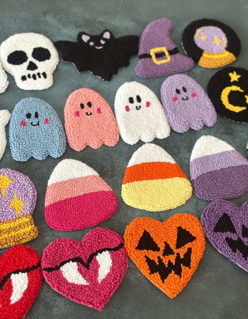 May include: A collection of handmade Halloween-themed coasters. Designs include a white skull, a black bat, a purple witch hat, ghosts, candy corn, and jack-o'-lanterns. The coasters are made of textured, colorful yarn.